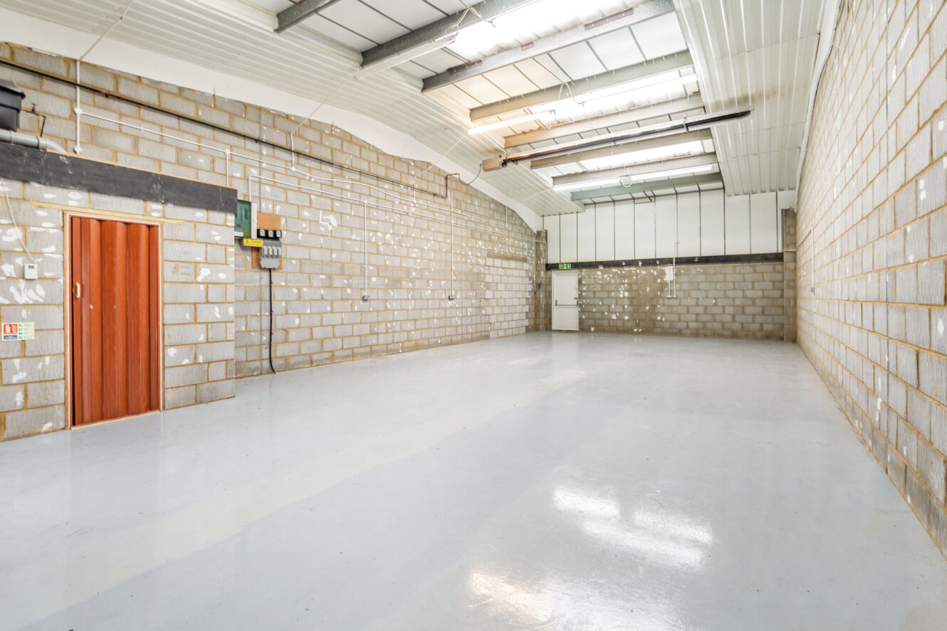 Empty warehouse with white floors, brick walls, a red door, fluorescent lighting, and exposed ceiling beams. Natural light enters through windows in the roof.