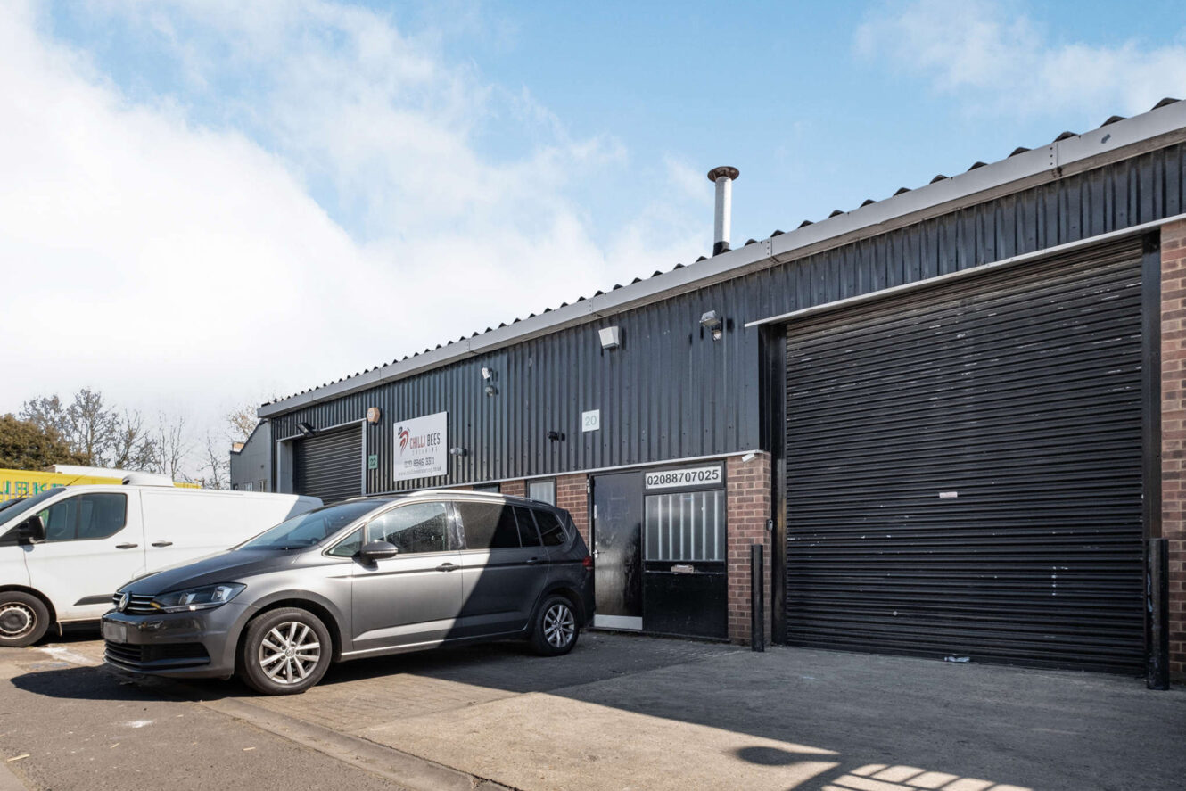 A black warehouse unit with a roller shutter door, a metal entry door, and two parked vehicles in front on a sunny day.