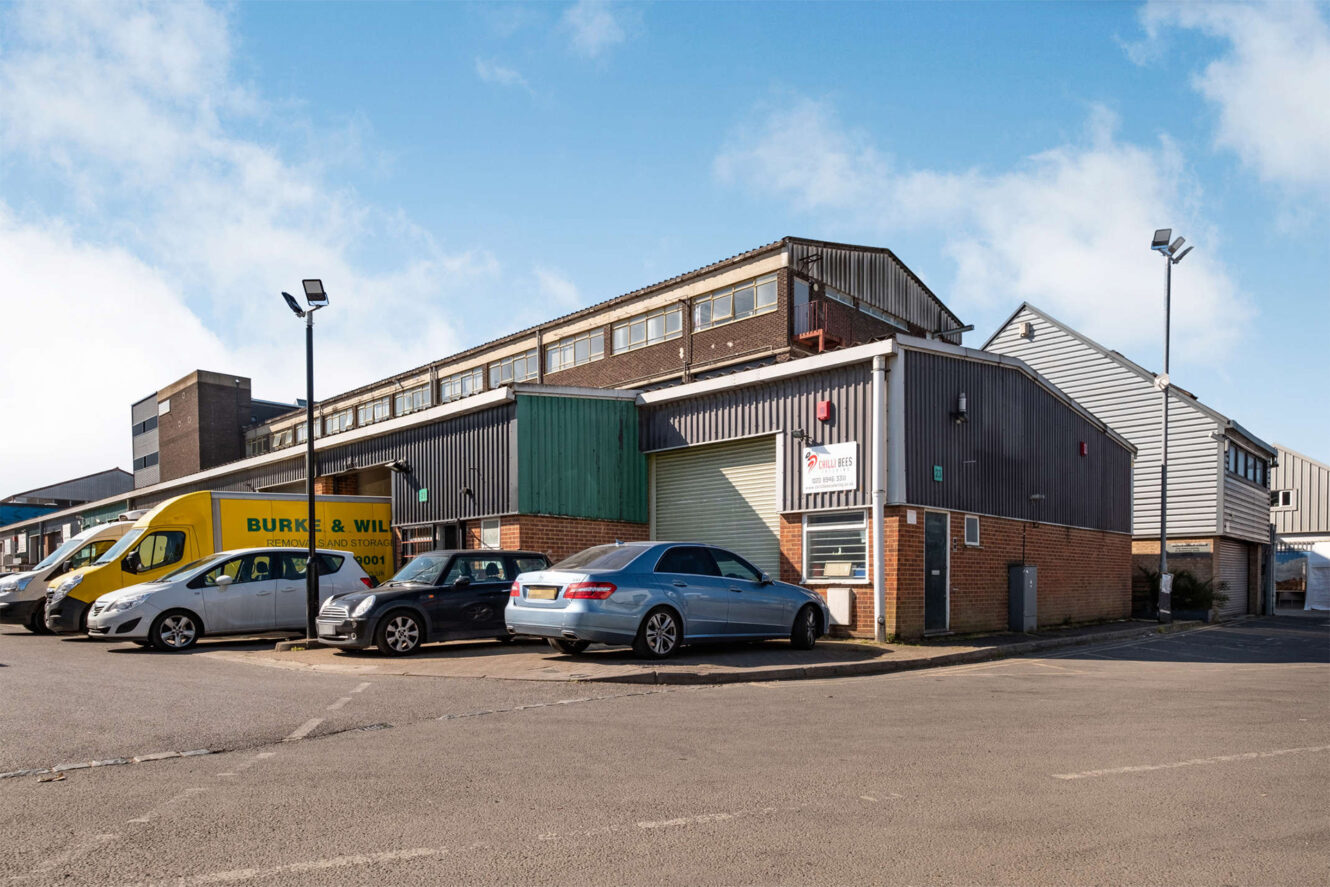A commercial warehouse building with several parked cars and a yellow delivery van visible in the parking lot under a clear blue sky.
