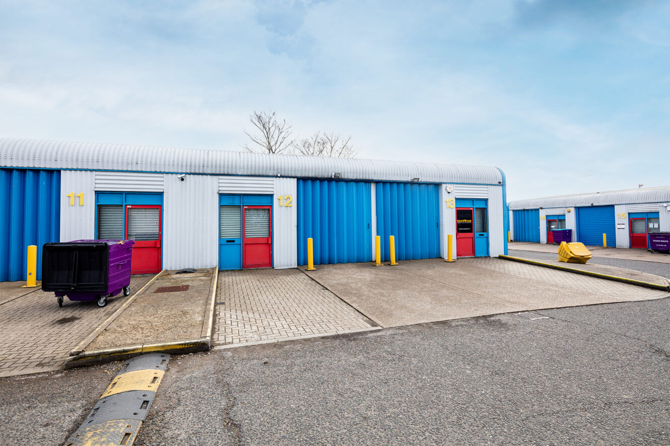 A row of industrial storage units with blue and white exteriors, numbered 11 to 13, each with red doors and yellow bollards; a purple bin is in front of unit 11.