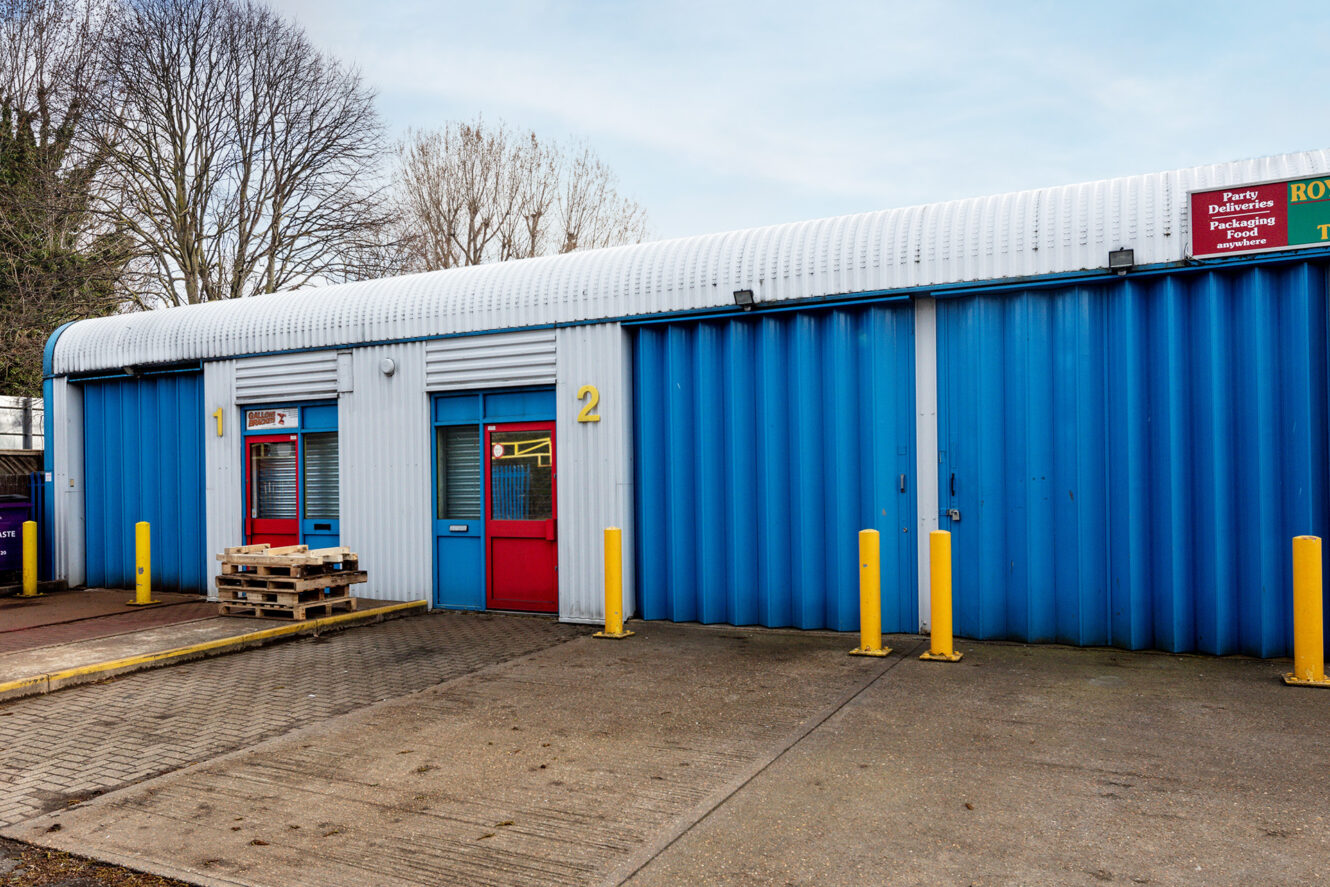 A blue and white industrial building with red doors, numbered 1 and 2, yellow bollards, and wooden pallets stacked outside.