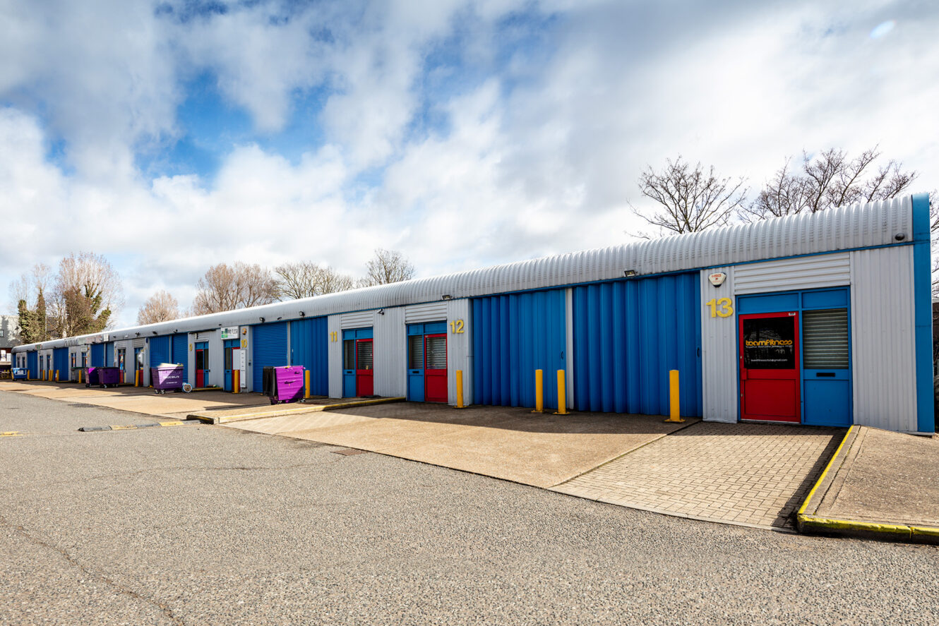 Row of blue storage units with red doors and numbered signs, set under a partly cloudy sky in a paved outdoor area.