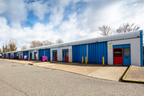 Row of blue industrial storage units with numbered red and gray doors, surrounded by pavement and a partly cloudy sky overhead.