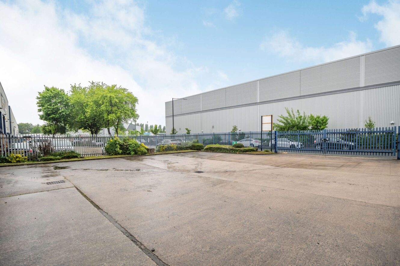Empty industrial yard with concrete ground, metal fence, some greenery, and large warehouse building in the background under a partly cloudy sky.