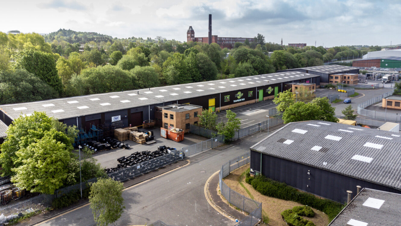 Aerial view of an industrial estate with large warehouses, office buildings, and surrounding trees under a partly cloudy sky.