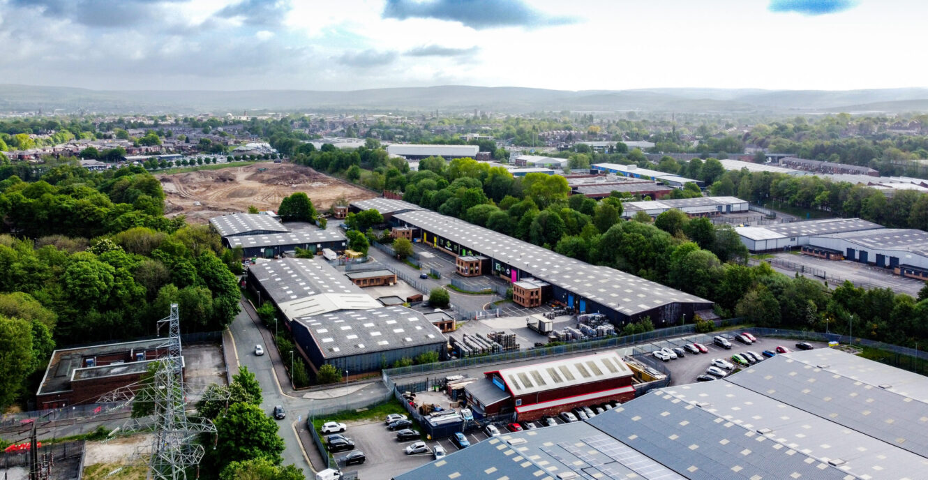 Aerial view of an industrial estate with several large warehouse buildings, parking lots, surrounding trees, and distant residential areas under a cloudy sky.