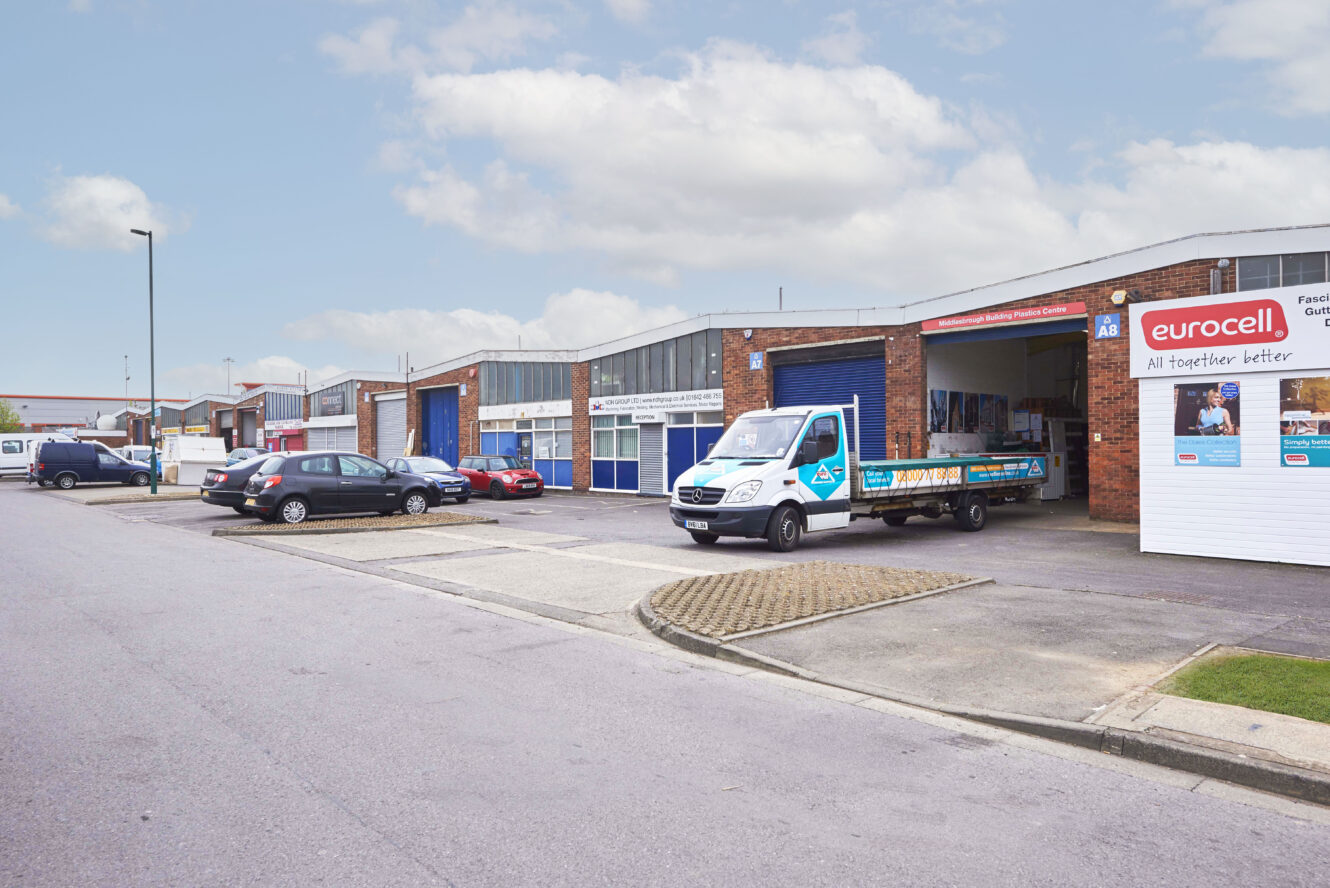 A row of industrial units with blue doors; cars are parked outside, and a flatbed delivery truck is parked in front of a Eurocell storefront.