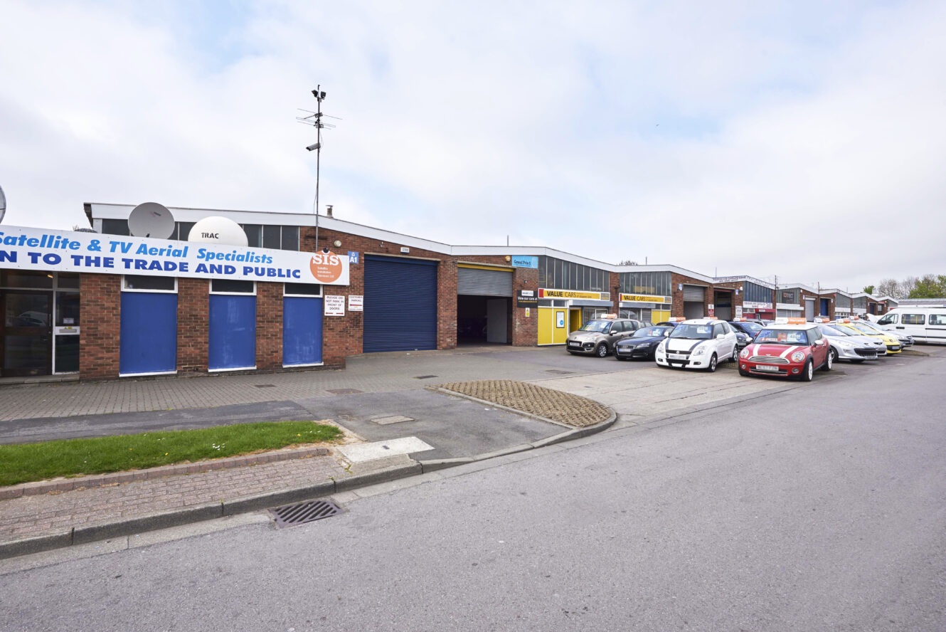 A row of single-story commercial units with various signs and parked cars along the street in an industrial area.