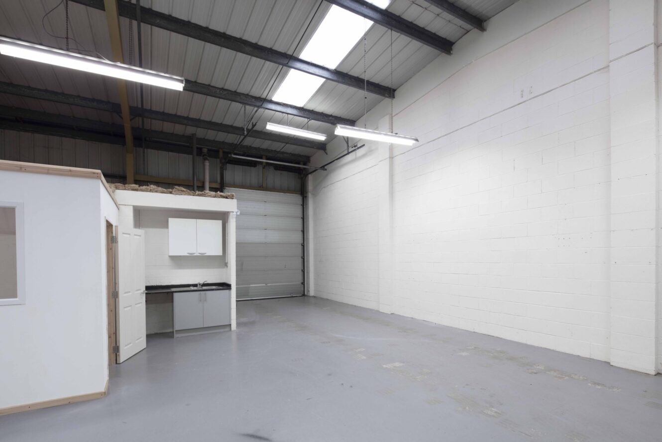 Empty industrial warehouse space with a small kitchenette area, white walls, high ceiling, large overhead lights, and exposed metal beams.