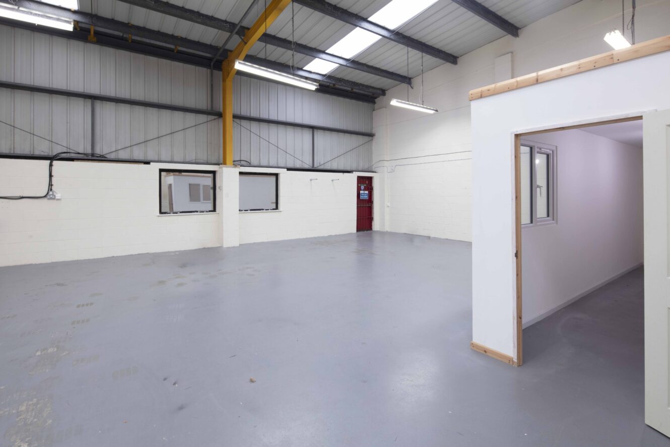 Empty industrial warehouse interior with gray concrete floor, white walls, exposed yellow beam, overhead lighting, and a small enclosed office room with windows on the right.