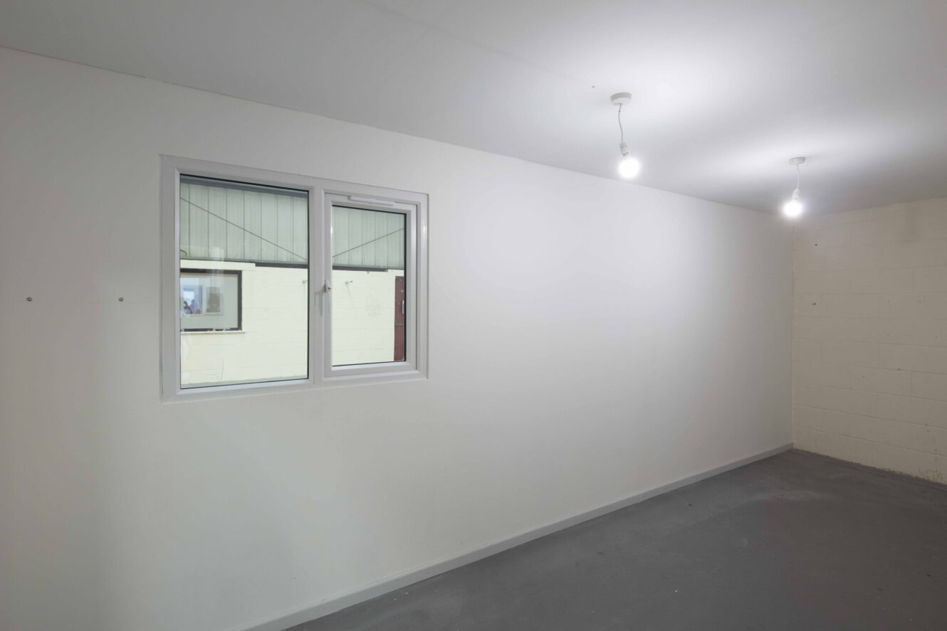 Empty room with plain white walls, two ceiling light bulbs, a window on the left wall, and a gray concrete floor.