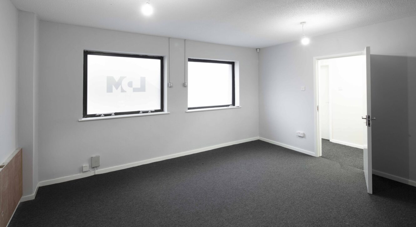 Empty room with white walls, dark carpeted floor, two windows, and an open door leading to a brightly lit adjacent room.