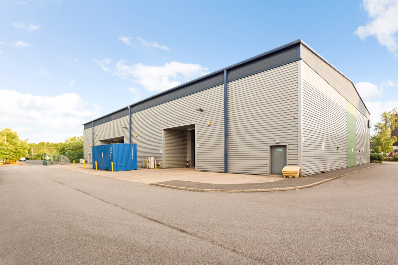 Large industrial warehouse building with metal siding, several loading bays, a blue shipping container, and a mostly empty paved lot under a clear sky.