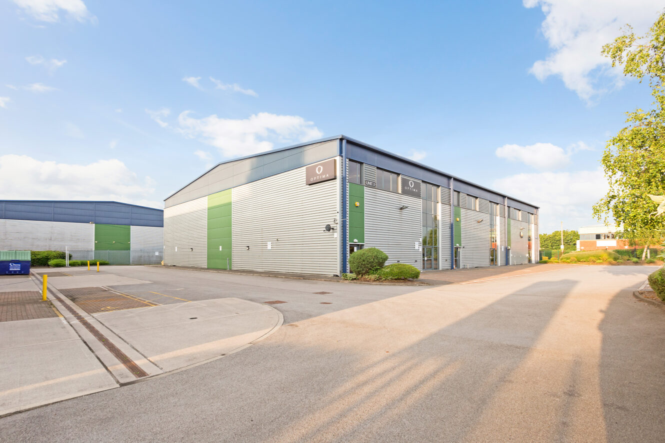 A modern industrial warehouse with metal siding, large green doors, and glass panels, located in a spacious, clean business park under a partly cloudy sky.