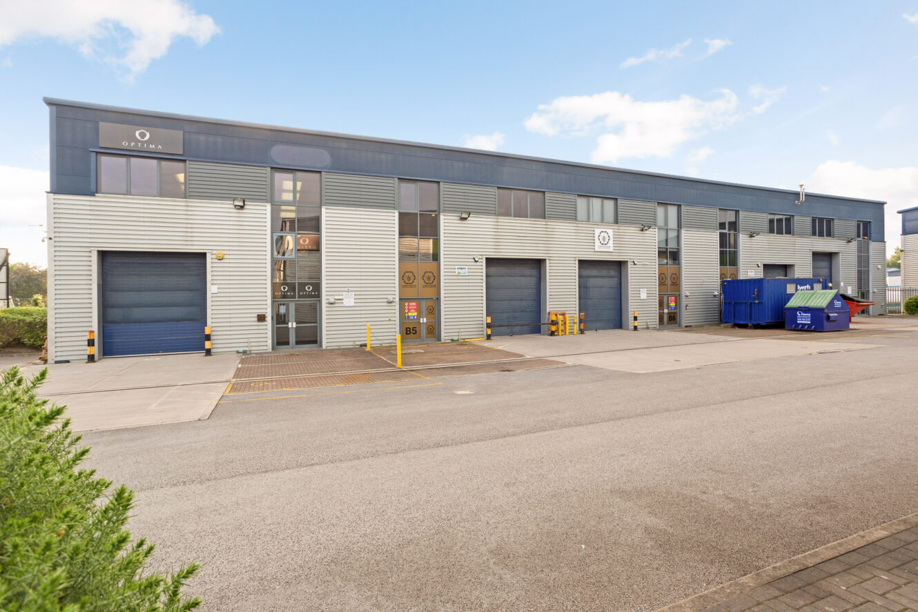 A modern industrial building with multiple garage doors, glass windows, and a blue dumpster in the parking lot under a partly cloudy sky.