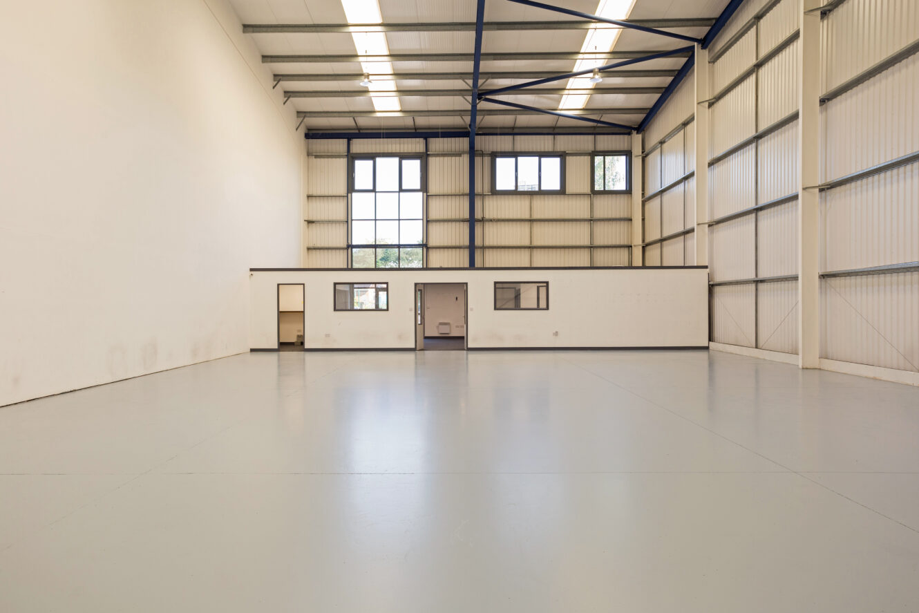 An empty industrial warehouse with high ceilings, large windows, exposed beams, and a small white office unit along the back wall.