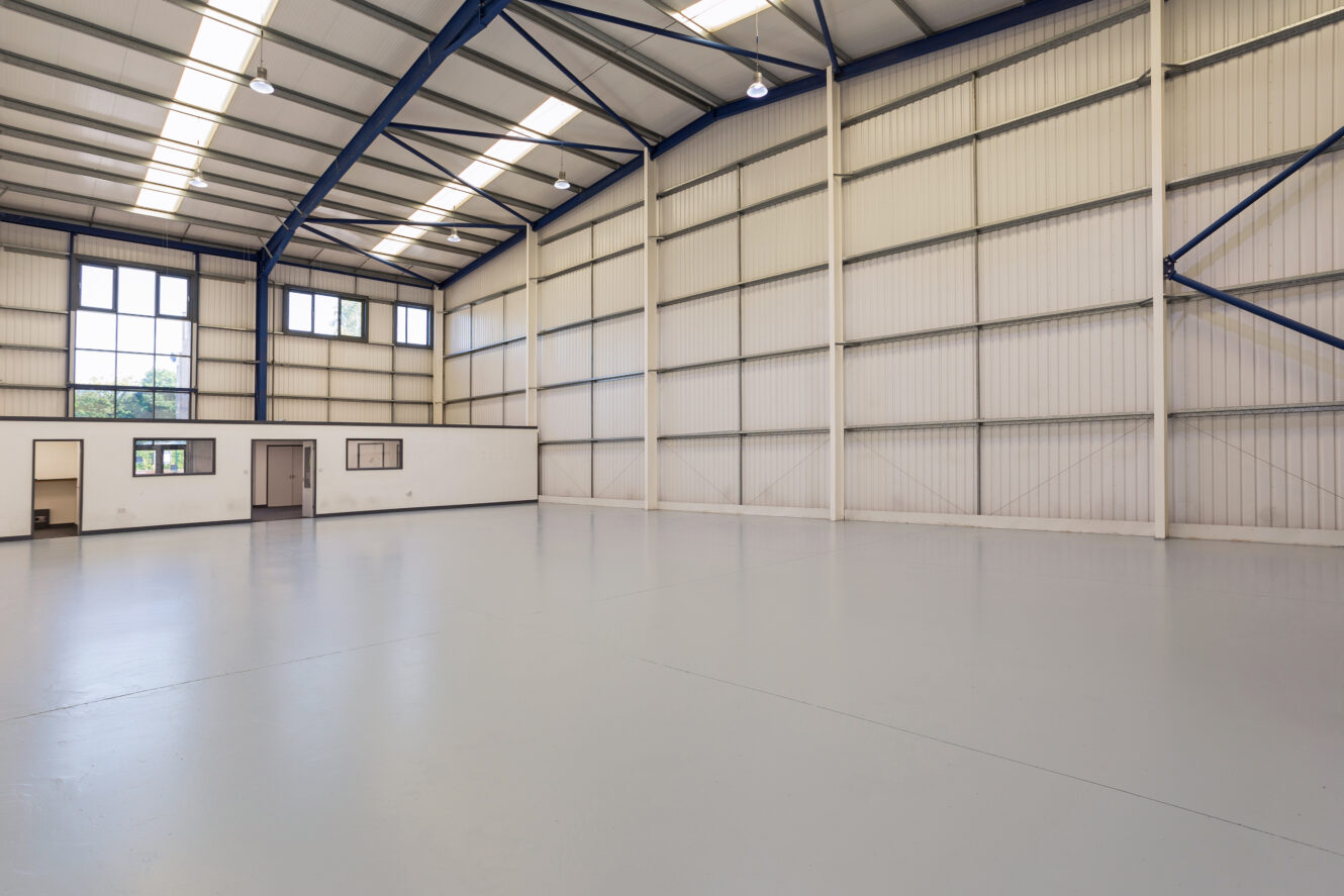 Interior of a large empty warehouse with high ceilings, metal beams, overhead lights, and an office area with windows and doors along one wall.