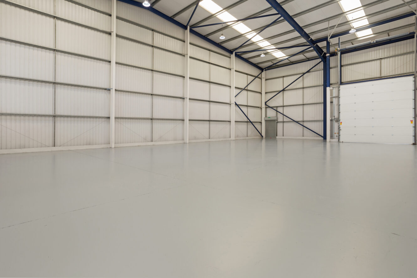 An empty industrial warehouse with a polished concrete floor, high metal walls, exposed beams, and a large closed roller door.