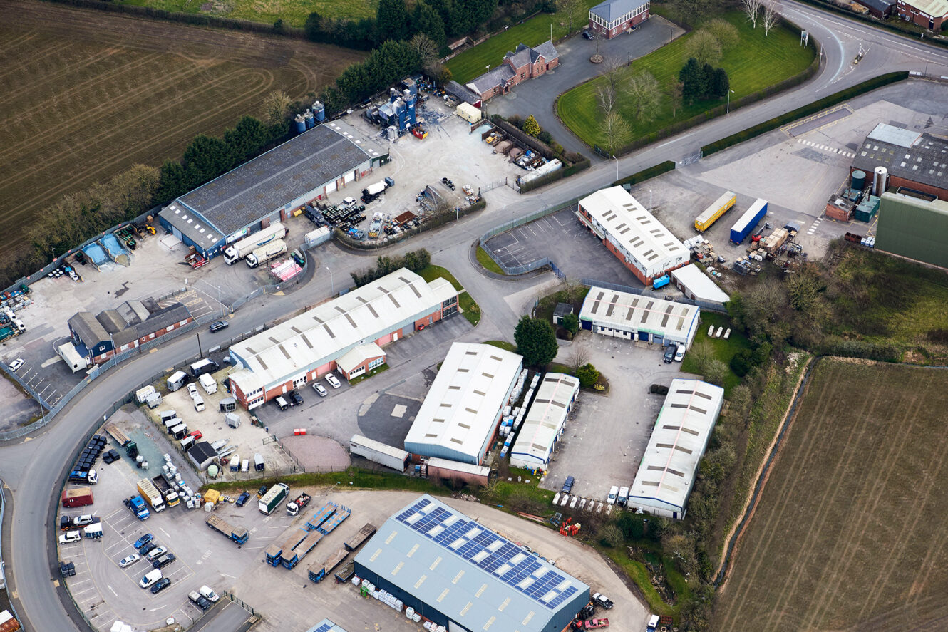 Aerial view of an industrial estate with several warehouses, parked vehicles, solar panels on one roof, and surrounding roads and fields.