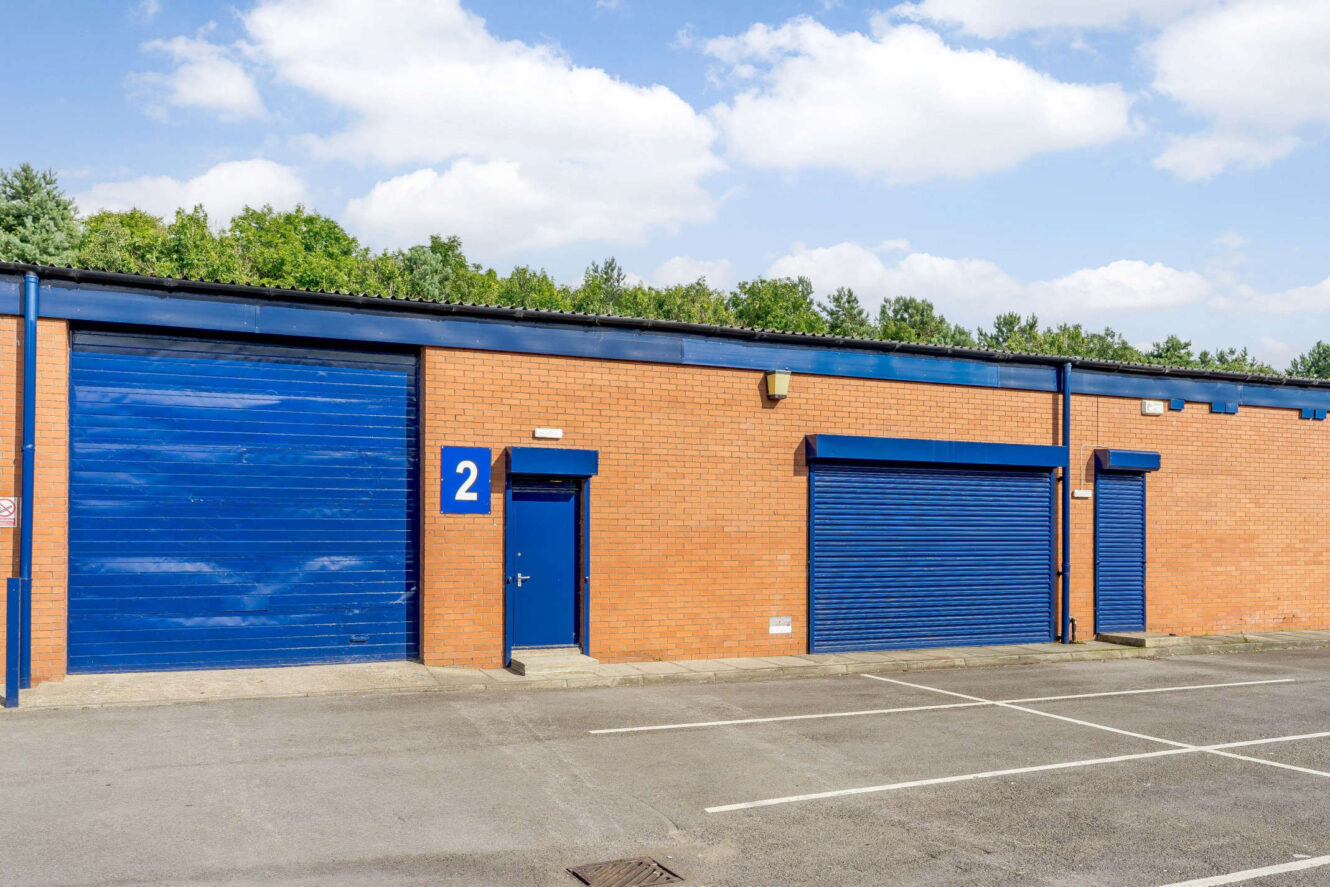 A brick industrial building with blue doors and shutters, marked with the number 2, along a row of parking spaces under a partly cloudy sky.