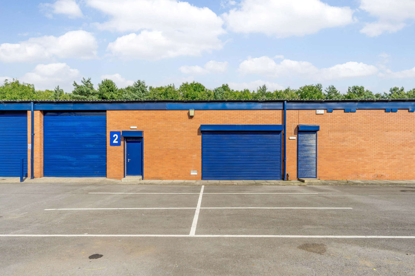 Brick building with blue doors and shutters, labeled with the number 2, in front of an empty parking lot; trees and sky in the background.