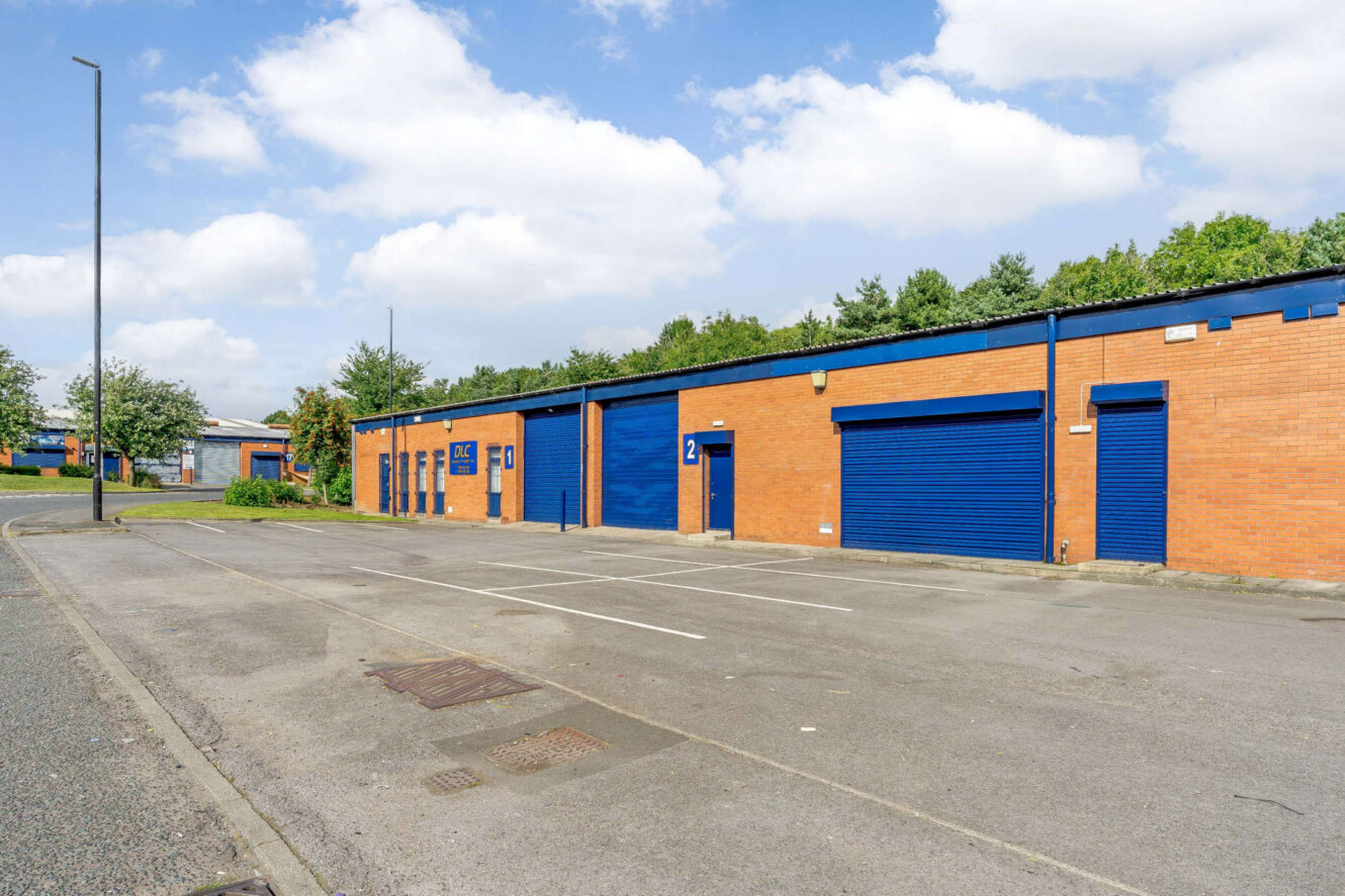 A row of industrial units with blue doors and shutters on a brick building, next to an empty parking lot and a street under a partly cloudy sky.