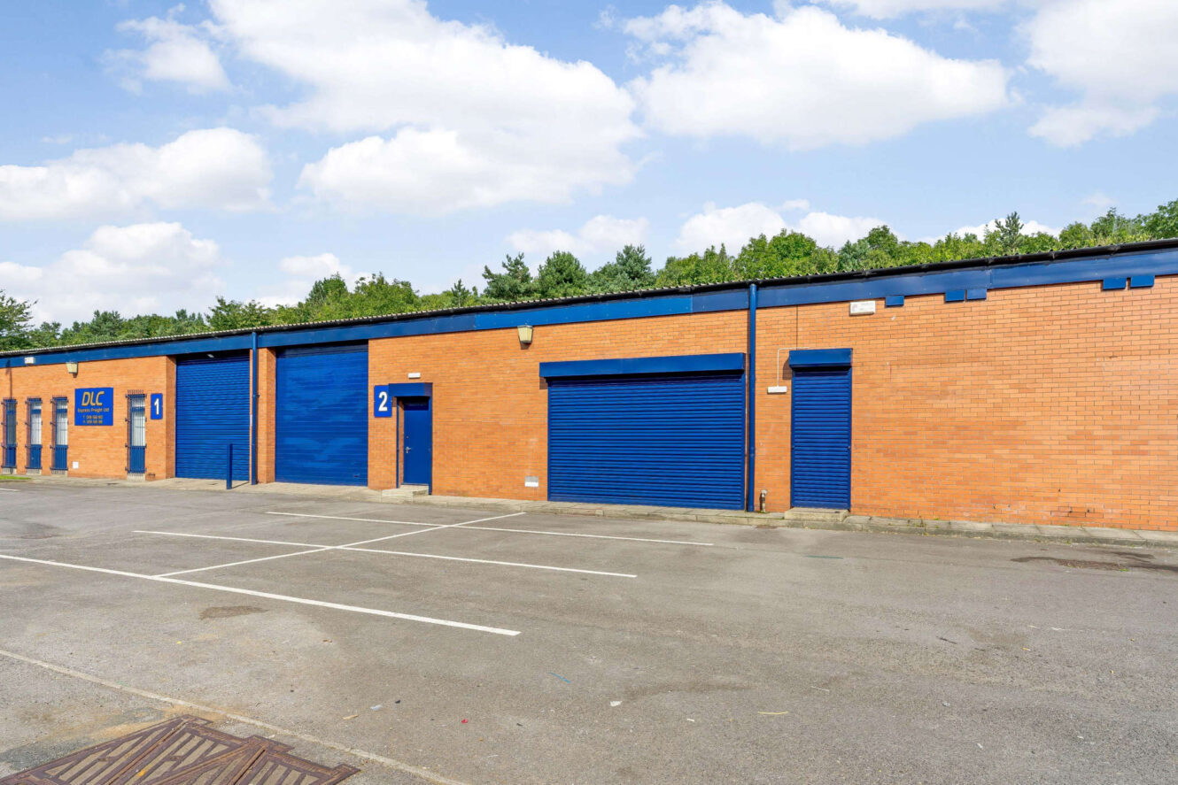 A row of brick industrial units with blue roller doors and doors, numbered 1 and 2, in front of an empty parking lot.