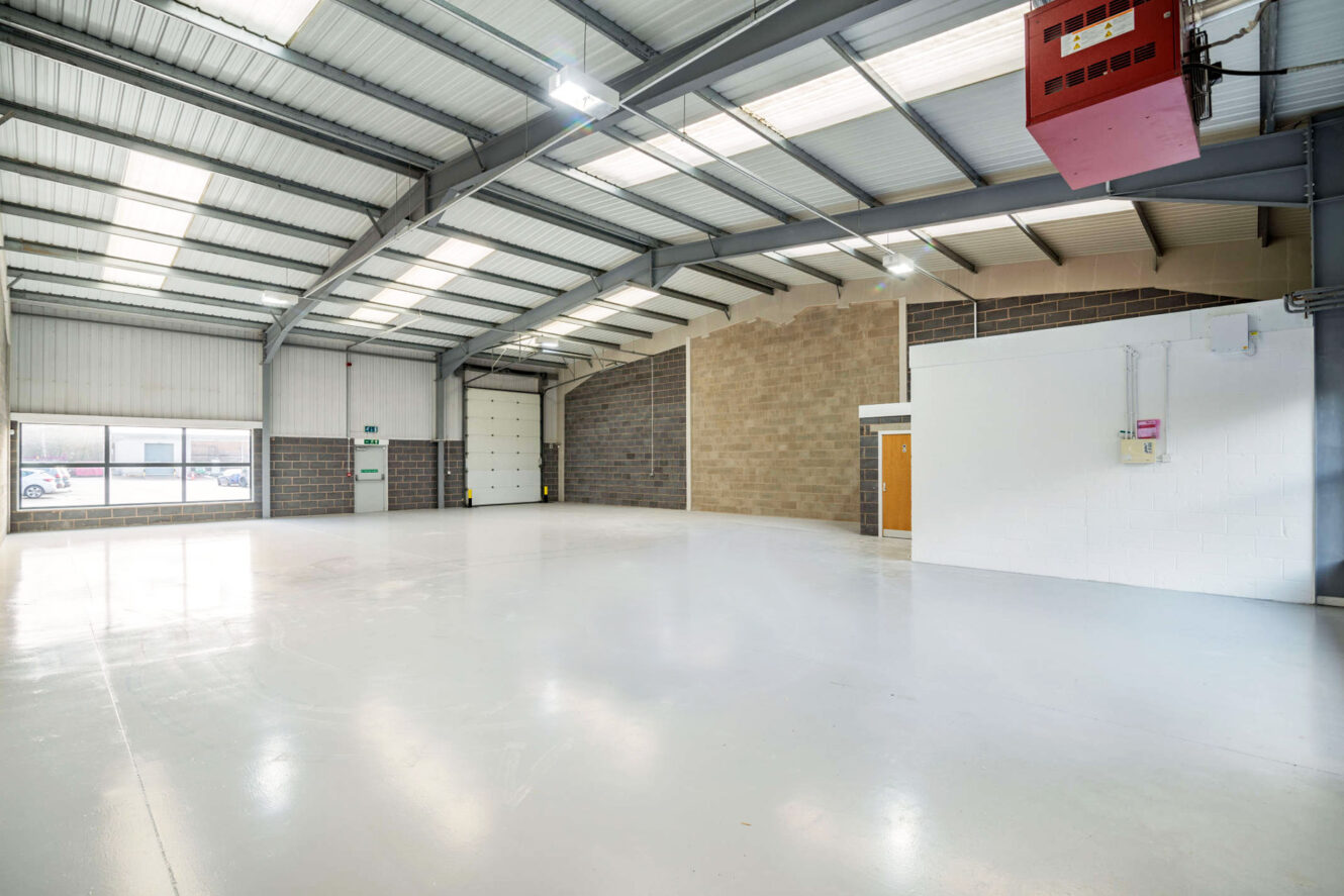 Interior of an empty industrial warehouse with white floors, brick and white walls, high ceilings, skylights, and a large garage door.