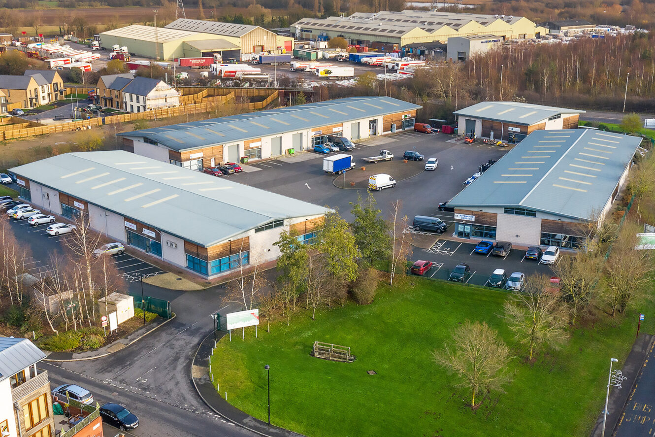 Aerial view of an industrial estate with several warehouses, parked vehicles, surrounding trees, and a nearby residential area in the background.