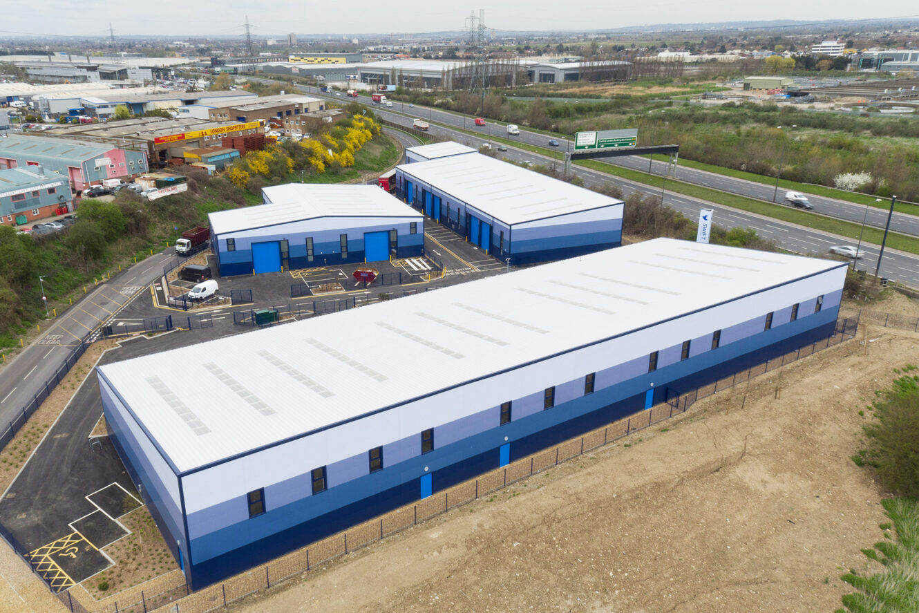 Aerial view of several blue industrial warehouse buildings next to a highway, with surrounding roads, parked vehicles, and nearby commercial properties.