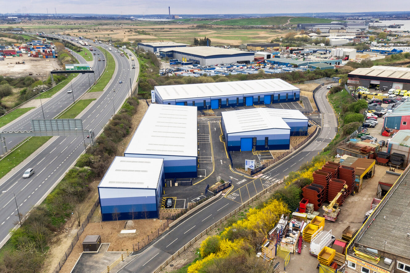 Aerial view of blue-roofed industrial buildings near a highway, with warehouses, vehicles, and construction materials surrounding the area.