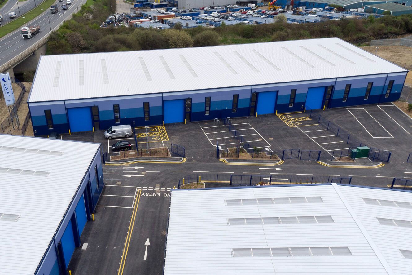 Aerial view of a modern industrial complex with several blue warehouse buildings, a parking lot with few cars, and surrounding fencing.