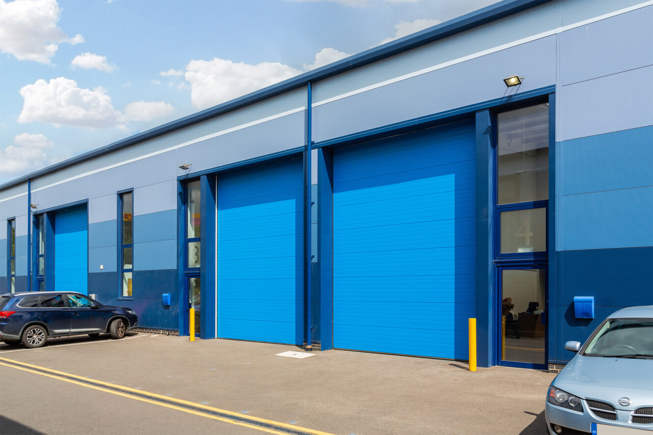 A row of blue industrial warehouse units with large roller shutter doors and parked vehicles outside on a sunny day.