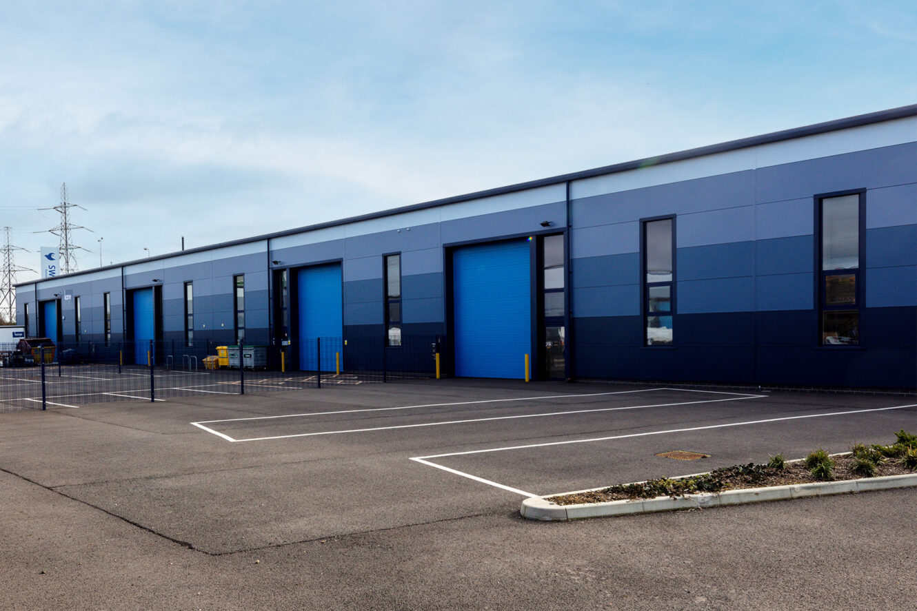 A modern industrial building with blue roller doors, large windows, and an empty parking lot in front, under a cloudy sky.