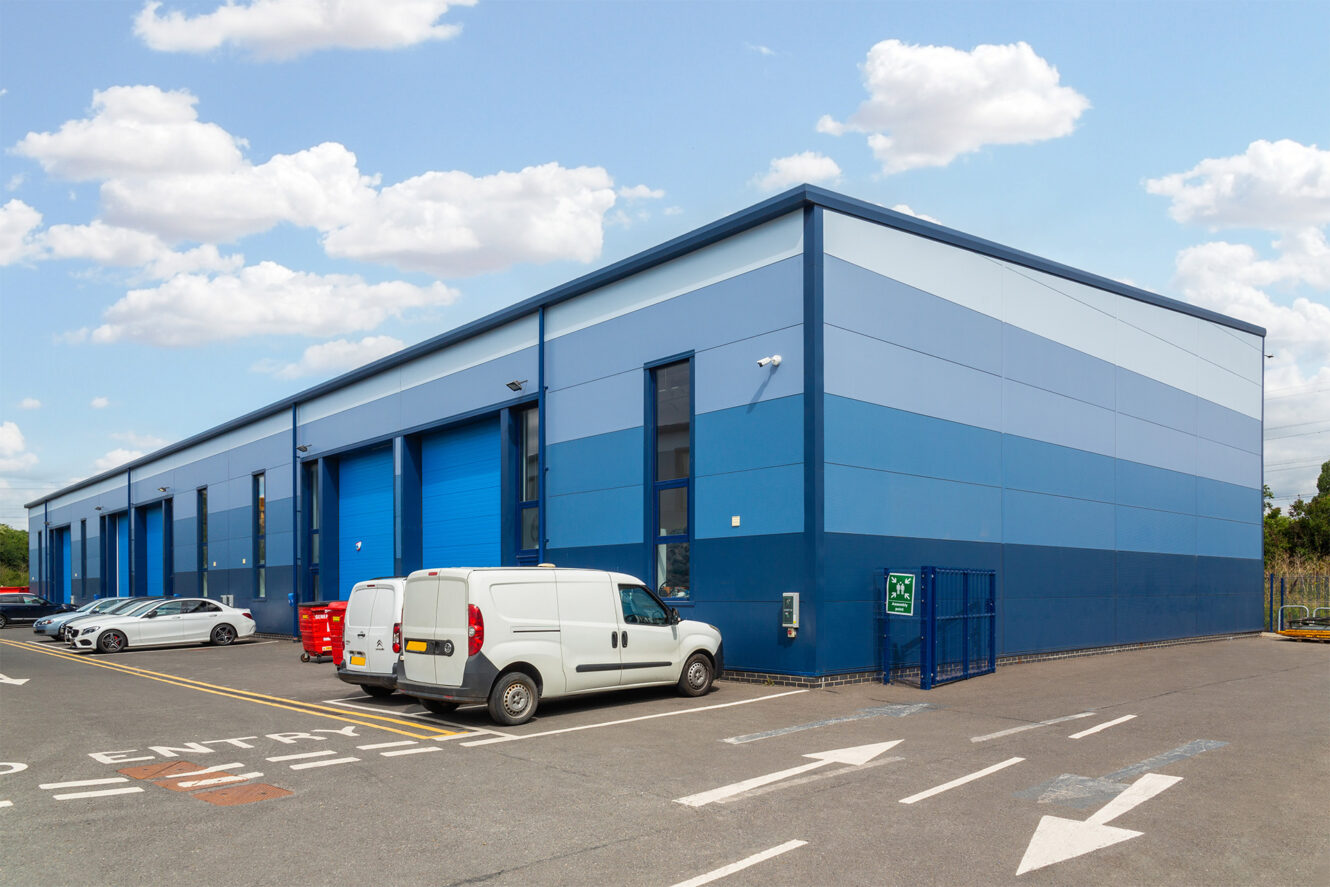 A modern industrial warehouse with blue paneling, several parked vehicles, and clear skies.