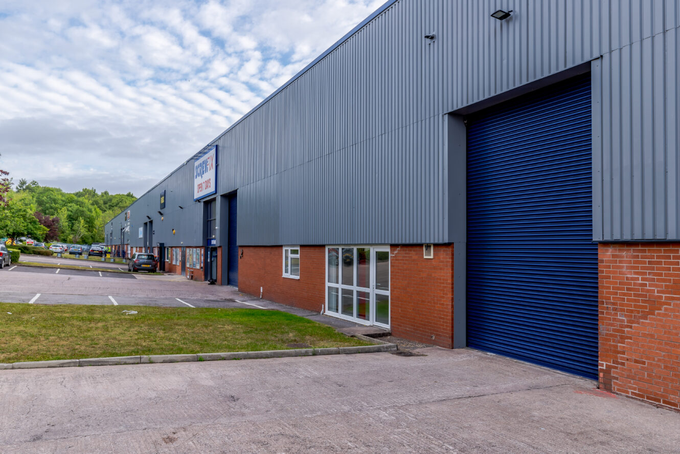 Large industrial warehouse with blue metal siding, a blue roll-up door, red brick lower walls, and a small parking area in front.