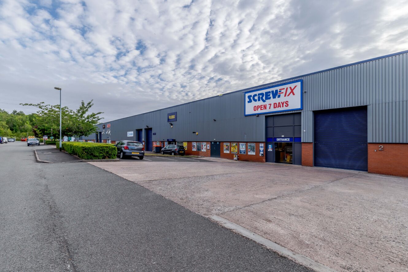 Large retail warehouse building with a Screwfix sign reading Open 7 Days, parking area in front, several cars parked, and a partly cloudy sky above.
