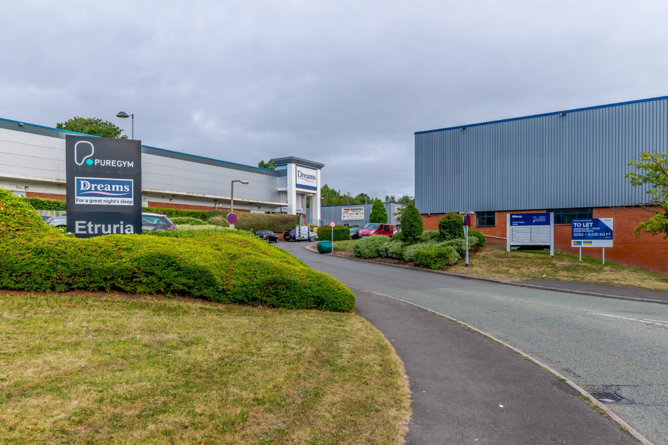A commercial area with signs for PureGym, Dreams, and Etruria; buildings and parked cars visible along a curved road with greenery and overcast sky.