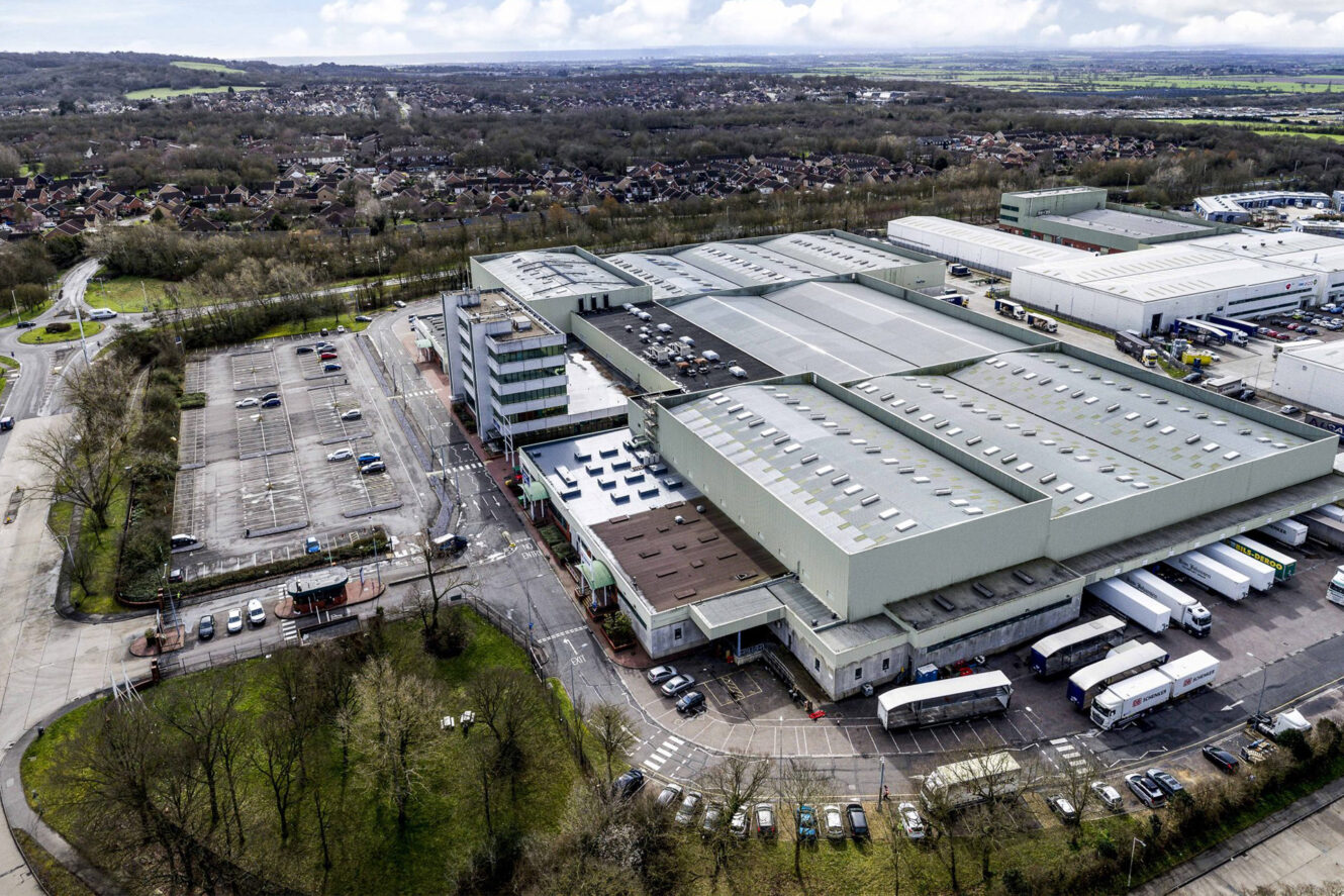 A large industrial warehouse with multiple loading docks, trucks, and an adjacent parking lot, surrounded by trees and residential areas in the distance.