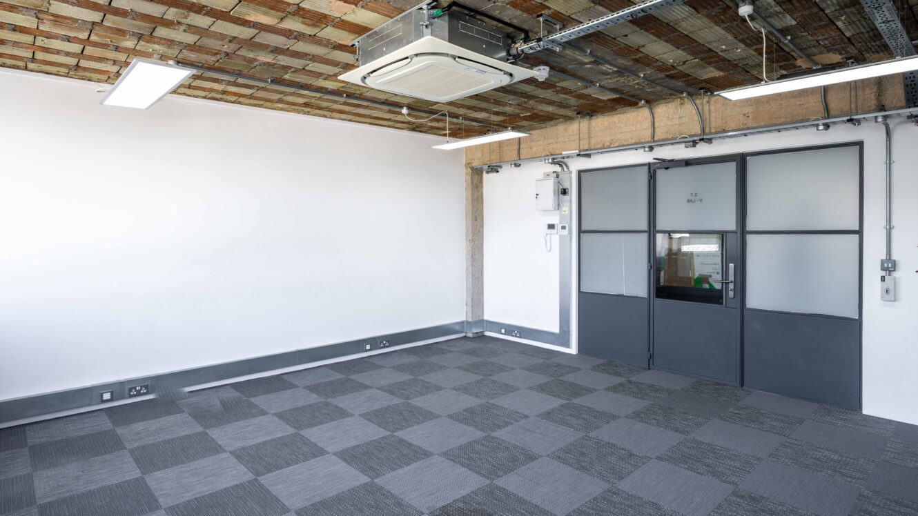 Empty modern office space with gray checkered carpet, white walls, exposed brick ceiling, and double glass doors.