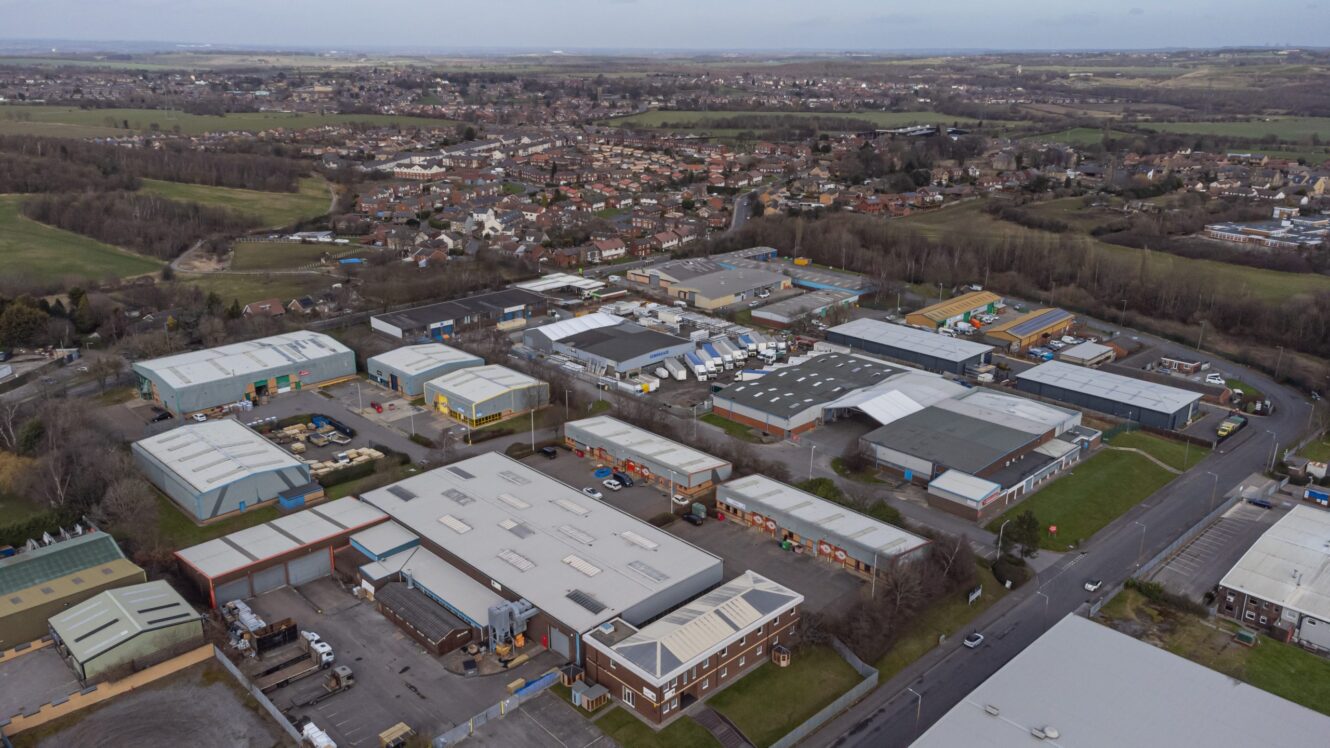 Aerial view of an industrial estate with multiple warehouses and office buildings, surrounded by roads and greenery, with a residential area in the background.