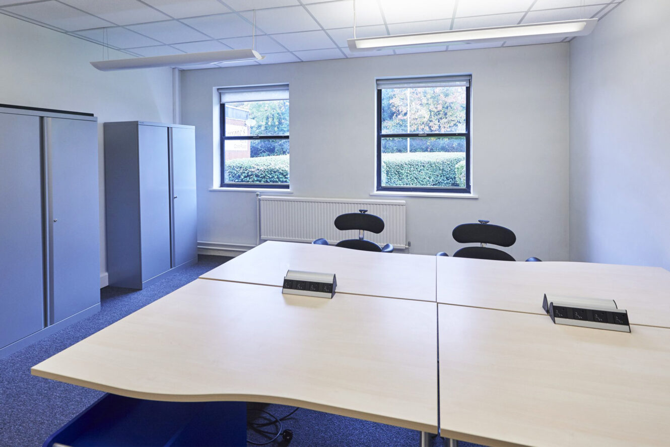 A small meeting room with four chairs around a square table, two windows, overhead lighting, and gray storage cabinets along the wall.