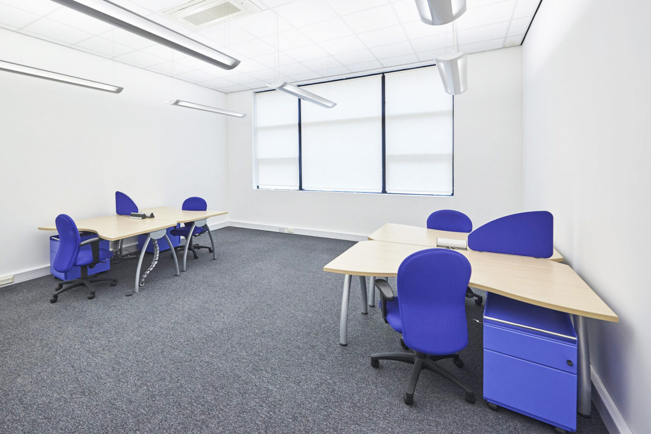 A modern office room with four desks, each with a blue chair and blue drawer unit, and large windows with blinds letting in natural light.