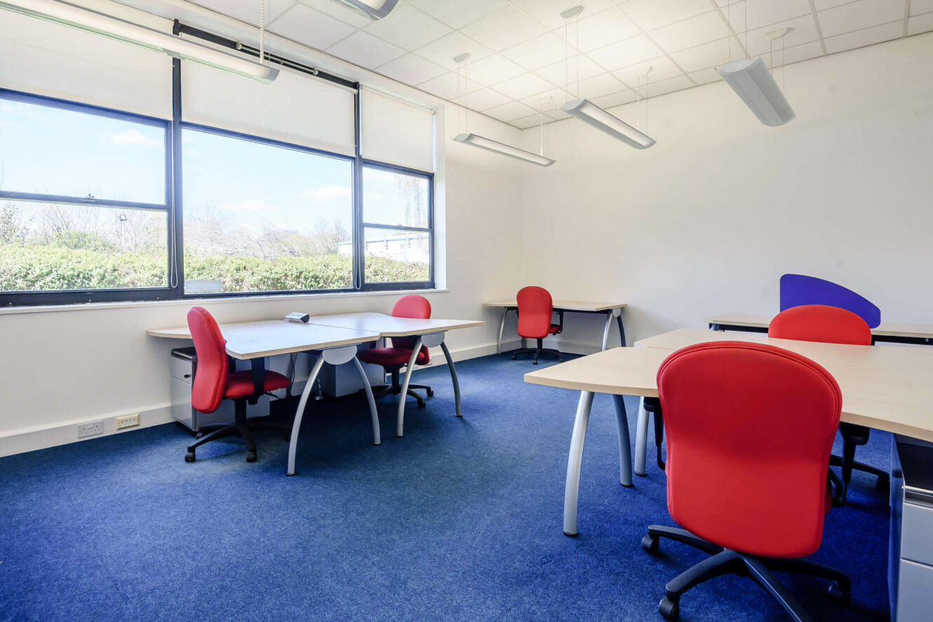 A modern office room with several desks and red chairs, large windows with natural light, blue carpet, and white walls.