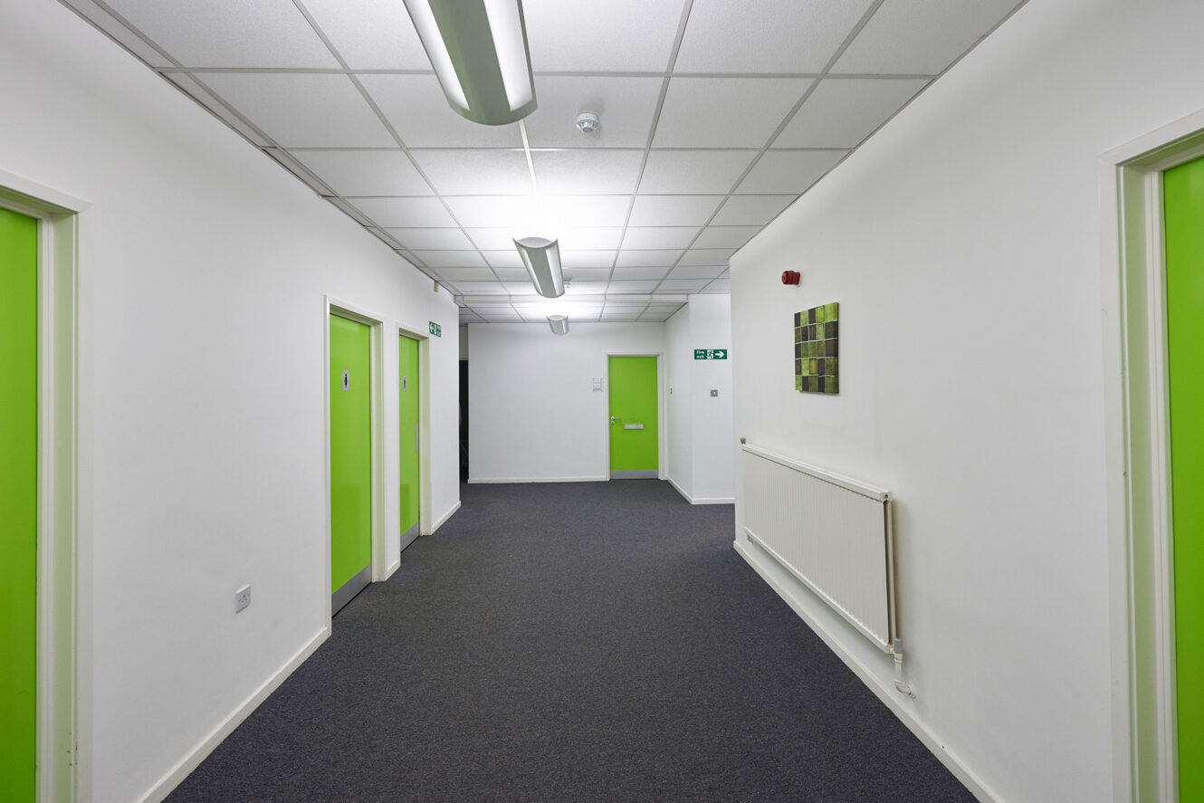 A clean, empty hallway with white walls, gray carpet, green doors, ceiling lights, and a wall-mounted radiator.