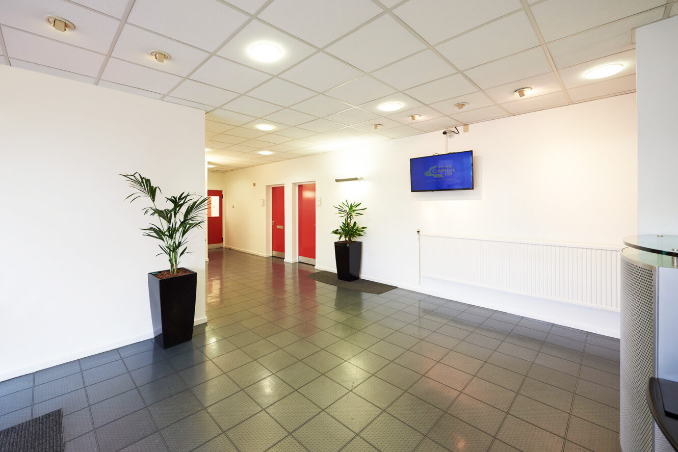 A clean, modern hallway with tiled floor, white walls, red doors, two potted plants, and a wall-mounted TV.