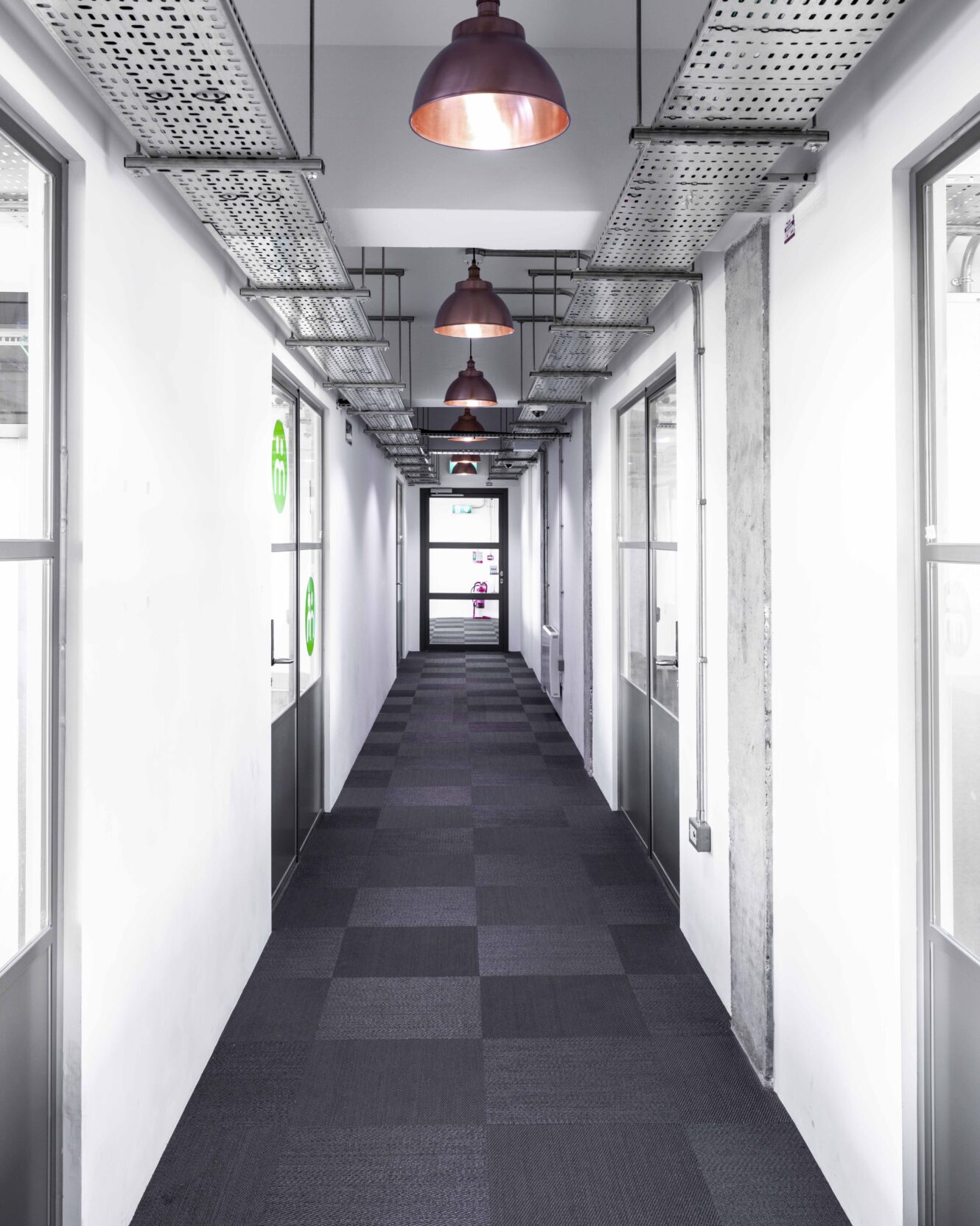 A modern, brightly lit hallway with grey carpet, white walls, glass office doors on both sides, and industrial-style ceiling lights.