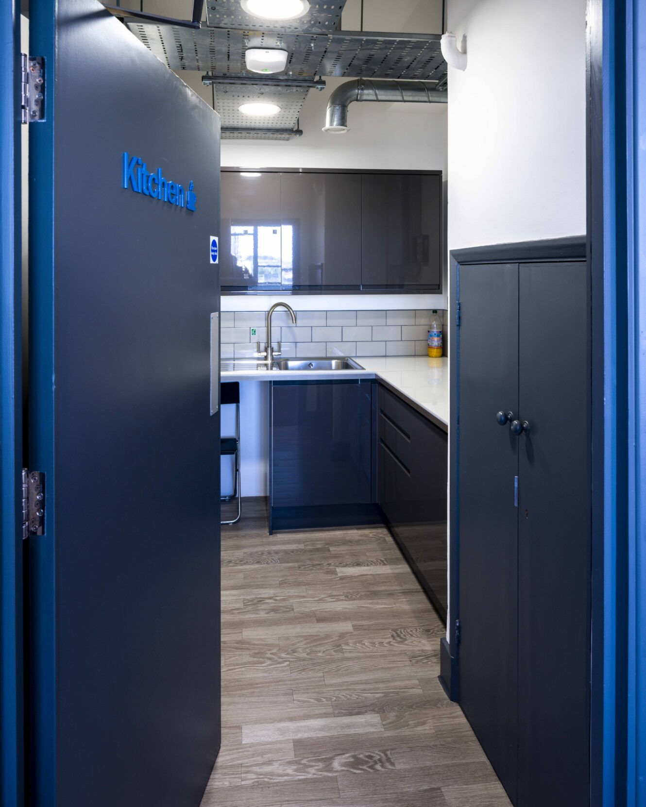Small modern kitchenette with dark cabinets, white countertop, sink, subway tile backsplash, and wood flooring, viewed through an open door labeled “Kitchen”.