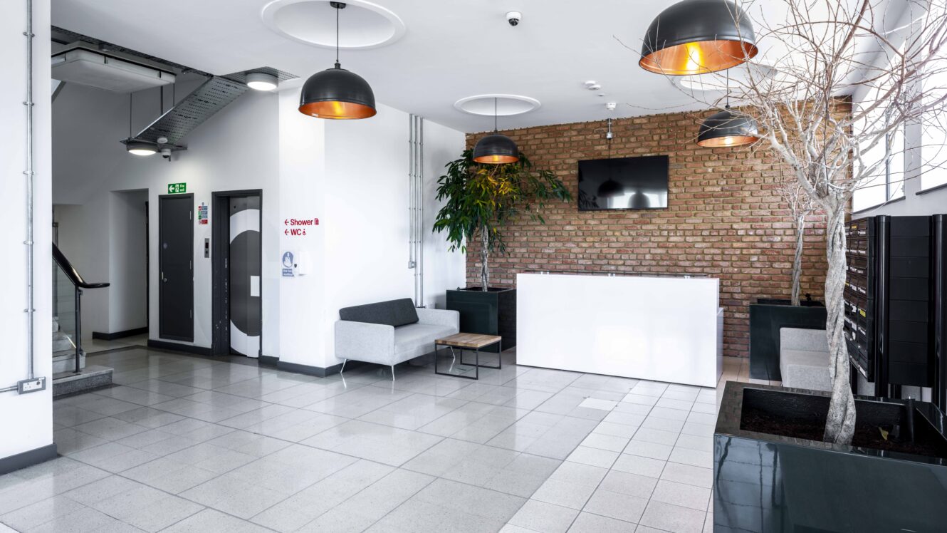 Modern office lobby with gray sofas, a white reception desk, brick accent wall, flat-screen TV, potted plants, and ceiling lights. Stairs and doors are visible to the left.