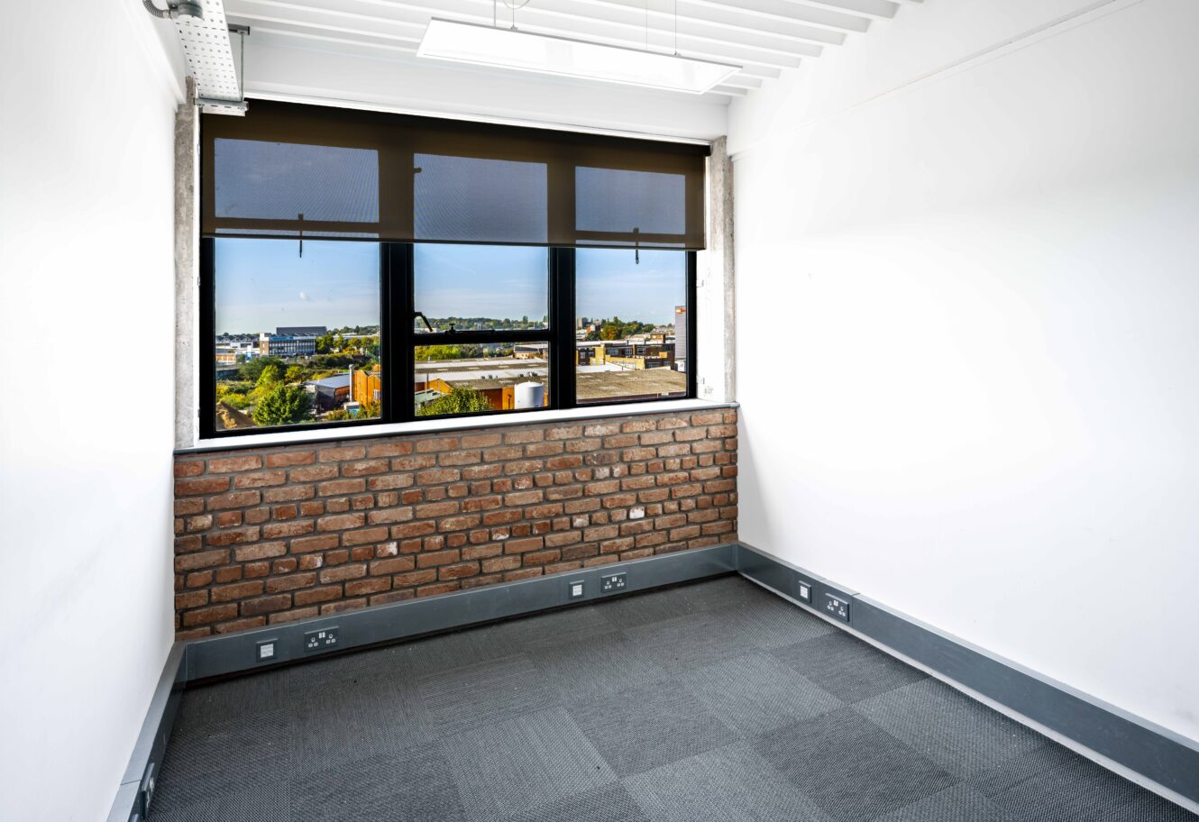 Small empty office room with brick accent wall, large window with black blinds, gray carpet, and multiple power outlets along the baseboard. Cityscape visible outside the window.